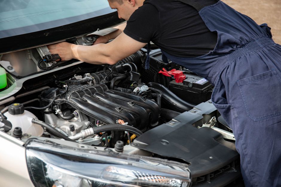 Mechanic in blue overalls working on car engine with open hood outdoors during the day.